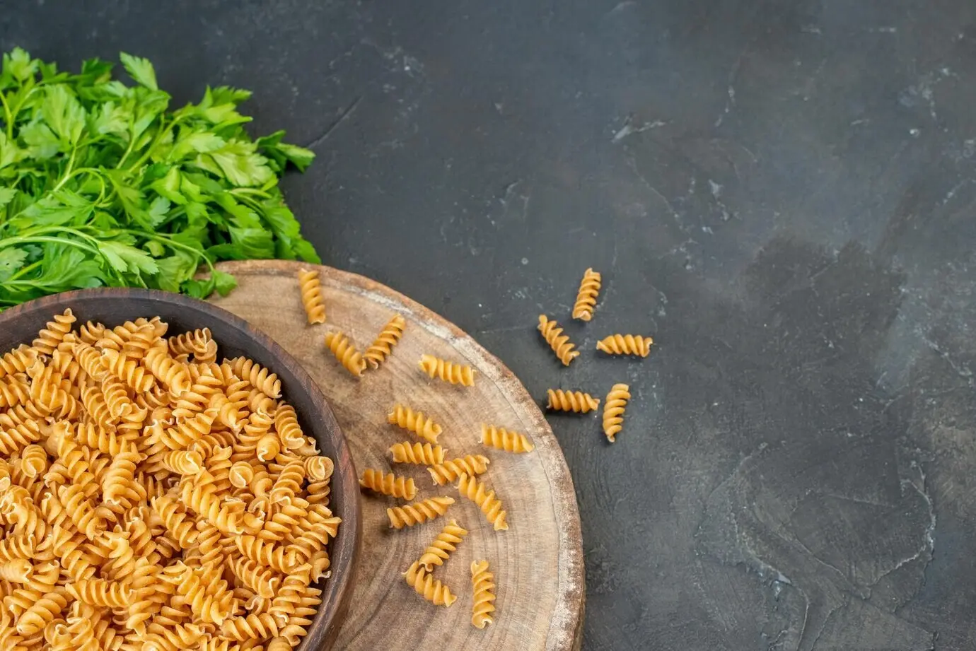 Top-down view of uncooked pasta inside and outside a brown pot, with a green bundle on the right side, on a dark blue background.
