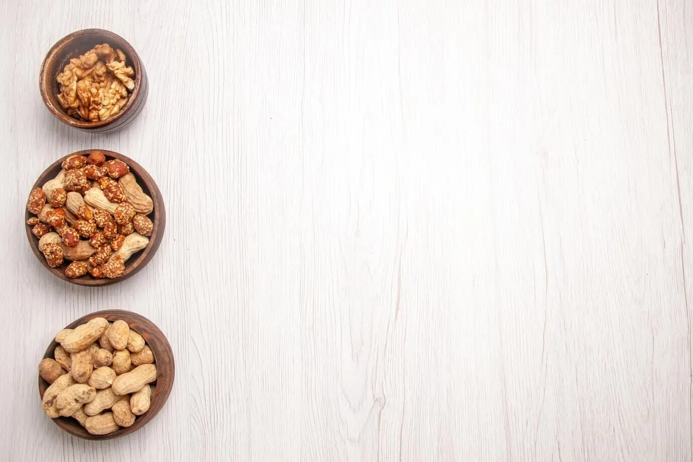 Overhead view from afar of three bowls of peanuts and walnuts on the left side of the white table.