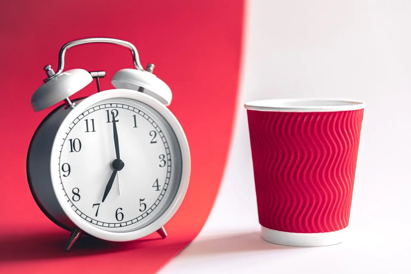 A morning concept featuring a white, stylish alarm clock and a paper cup.